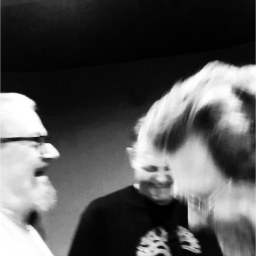 a black and white photo of the three band members laughing happily, one in the foreground on the right facing left, one in the midground on the left facing right and one in the background facing front looking down